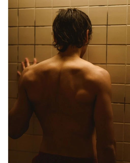 Back view of a light-skinned adult man leaning against a tiled wall indoors, lit in warm tones, showing his back and shoulders.