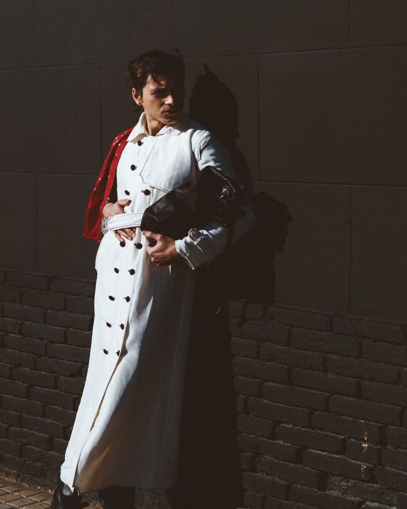 A fashionable person poses against a textured wall, wearing a white trench coat and clutching a black bag, lit by natural sunlight.