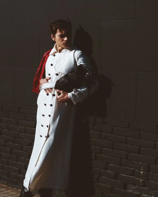 A fashionable person poses against a textured wall, wearing a white trench coat and clutching a black bag, lit by natural sunlight.