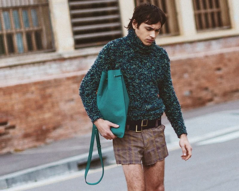 Male fashion model poses with green tote bag, wearing a textured sweater and plaid shorts outdoors with a building backdrop.