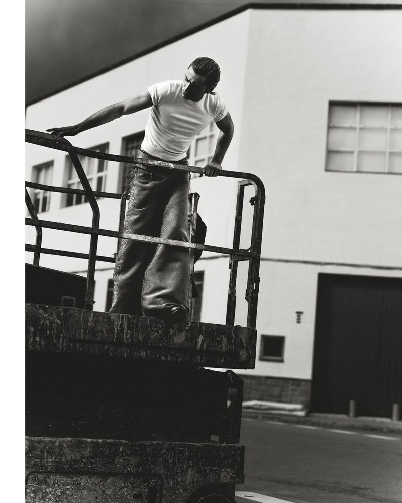 Monochrome portrait of a man in a white tee and wide-leg jeans leaning on a rusty metal structure, set against an industrial building.