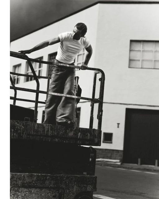 Monochrome portrait of a man in a white tee and wide-leg jeans leaning on a rusty metal structure, set against an industrial building.