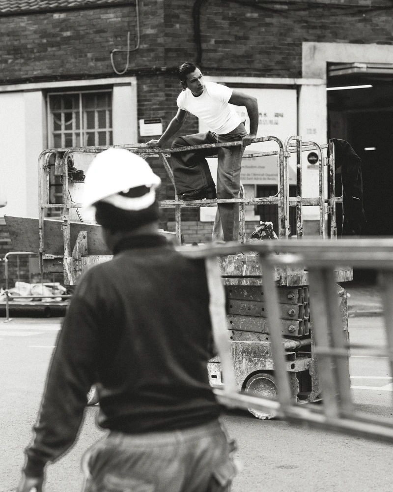 Black and white photo of a man in a white t-shirt on a scissor lift with a worker carrying a ladder in the foreground.