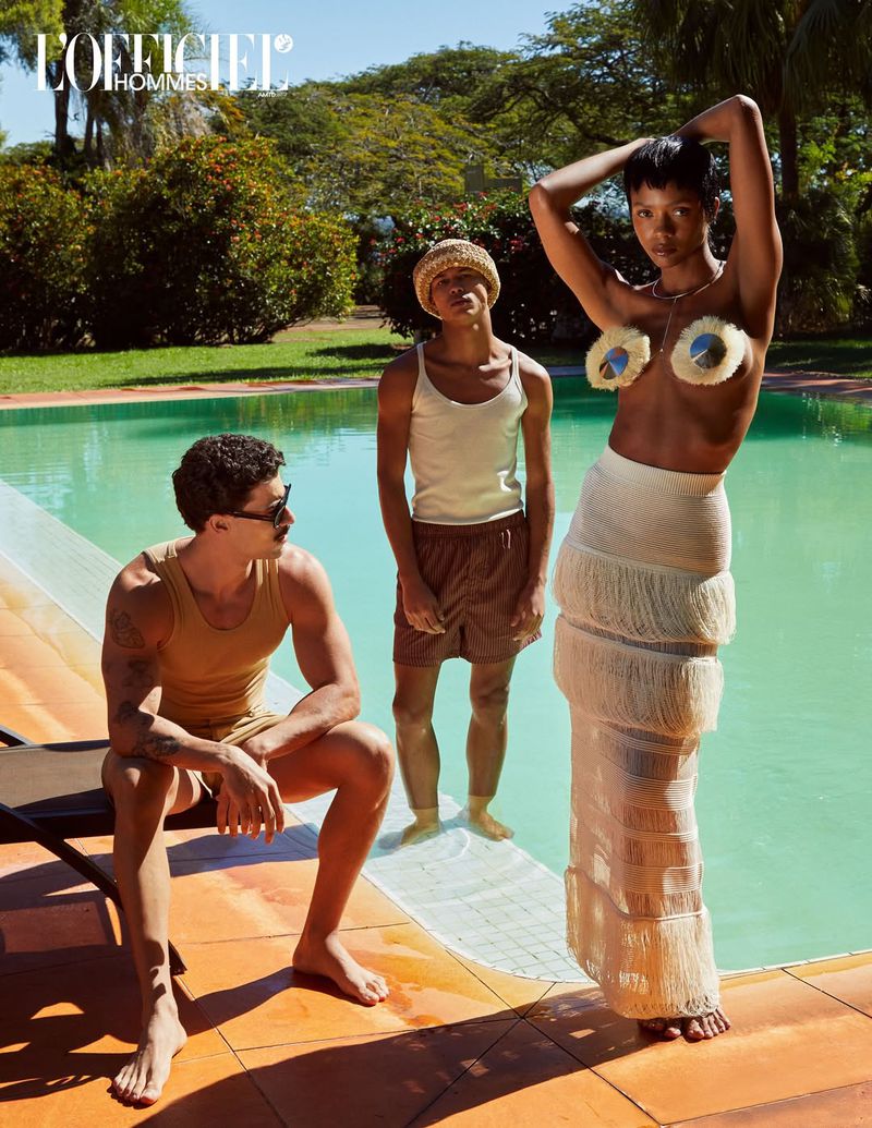 Three fashion models pose by a pool on a sunny day. The model on the right is wearing a white fringed skirt and decorative nipple covers.