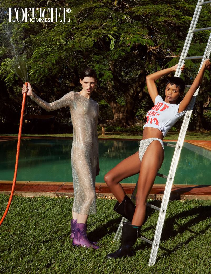 Two women pose outdoors on a lawn beside a ladder and pool, with one holding a hose and wearing a sparkling dress and purple boots.