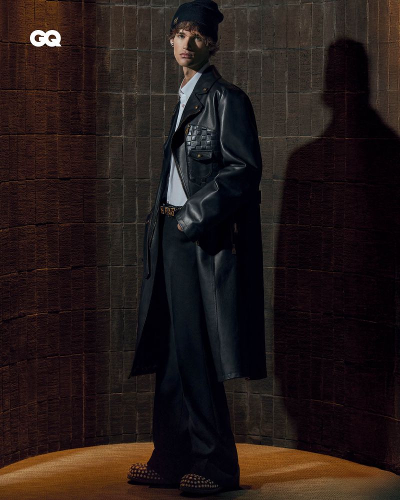 An adult male fashion model poses in a black leather trench coat, a white undershirt, and black pants with studded shoes.