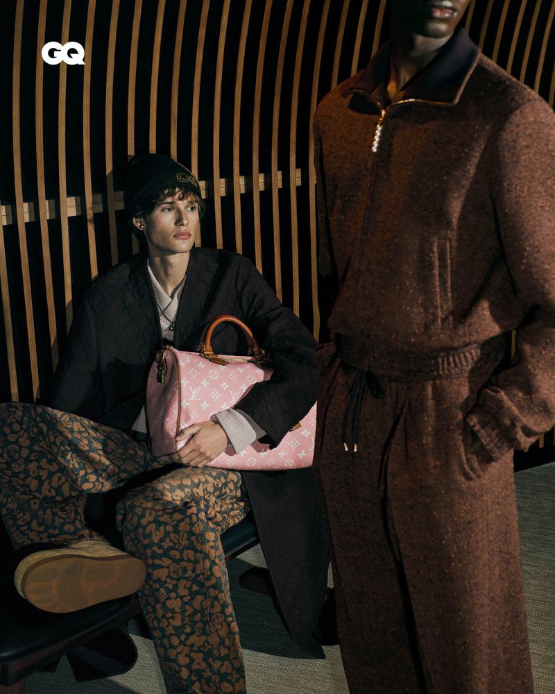 Editorial fashion shot of two male models indoors against a striped wall. One is seated with a Louis Vuitton bag, wearing patterned pants.
