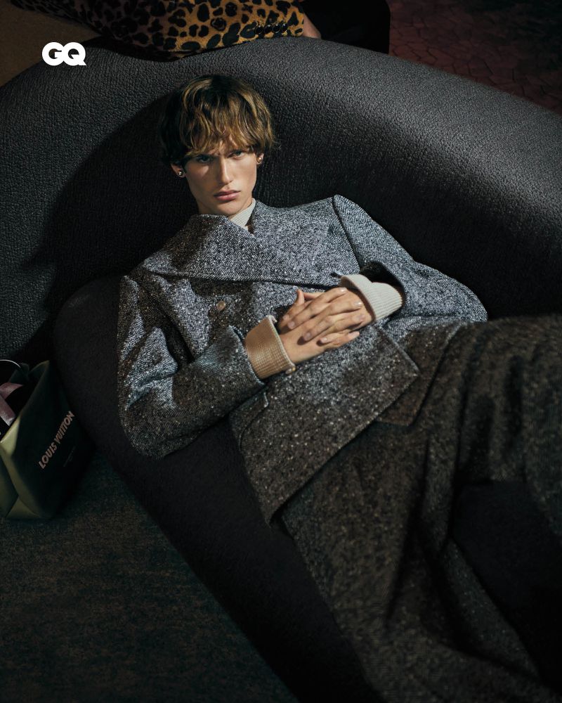 An adult is reclining on a gray couch, wearing a matching gray speckled jacket and pants, with a Louis Vuitton bag nearby and GQ logo.