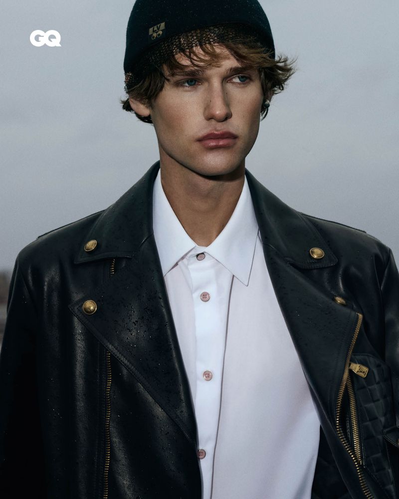 A young adult man poses wearing a black beanie, black leather jacket, and white collared shirt against an overcast sky.