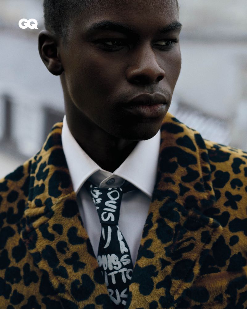 Portrait of a dark-skinned man wearing a cheetah-print coat and a patterned tie with a white shirt, photographed at eye-level.