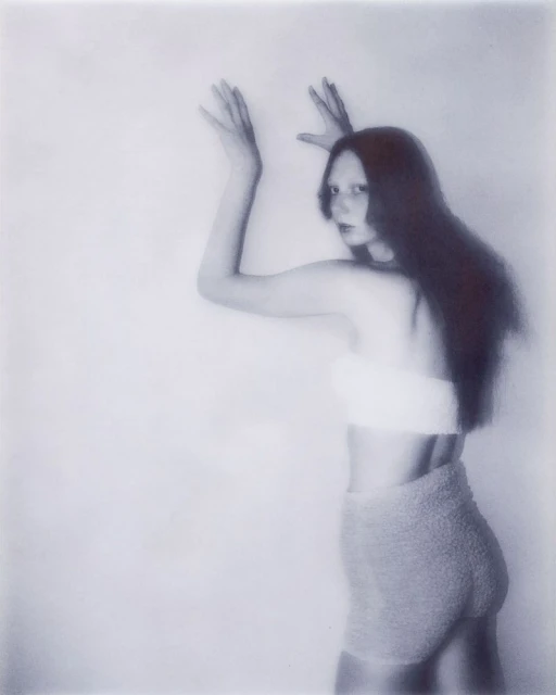 Woman with long dark hair poses in a light blue artistic photograph, wearing a white top and grey shorts, with her arms raised.
