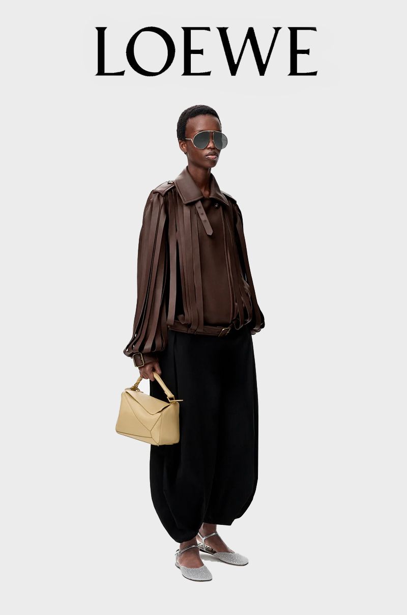 Fashion shot of a Black female model wearing a brown jacket and oversized black pants, holding a light-yellow handbag, in a studio.