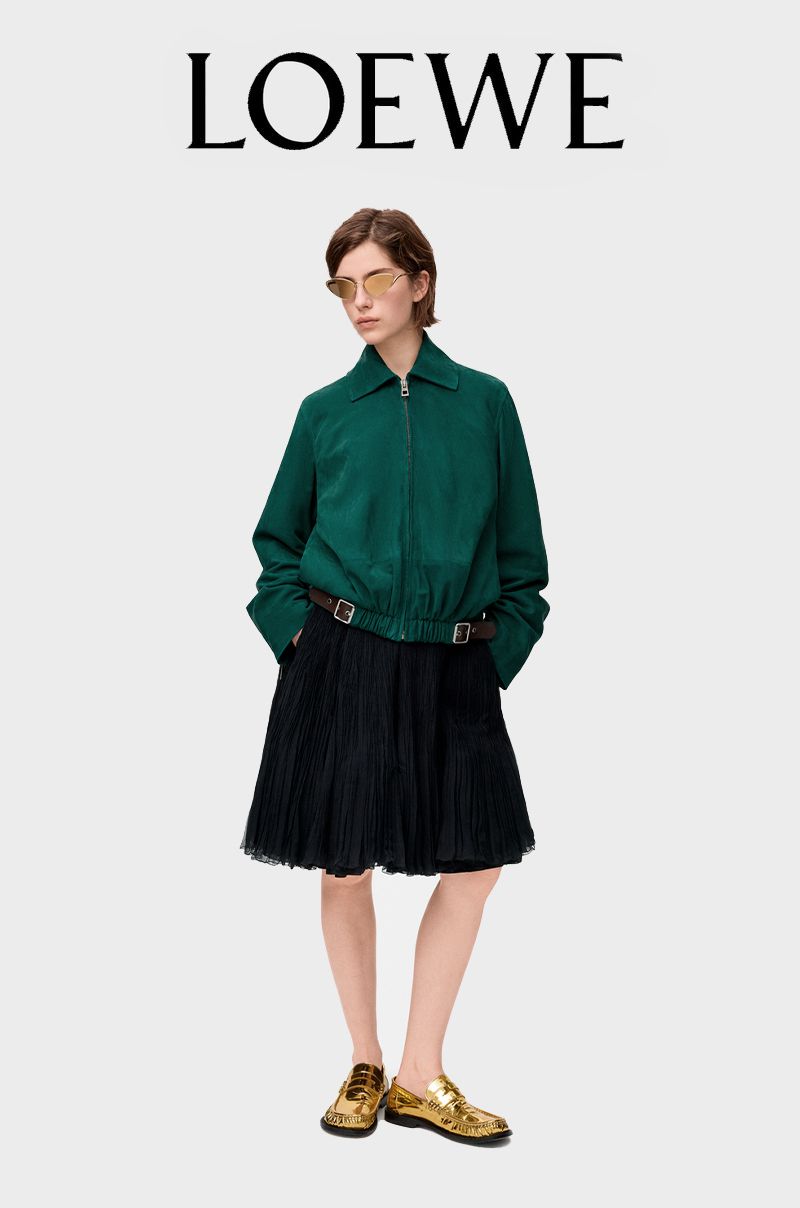 An adult woman poses in a green zip-up jacket, a black pleated skirt with a brown belt, and gold shoes on a grey backdrop.