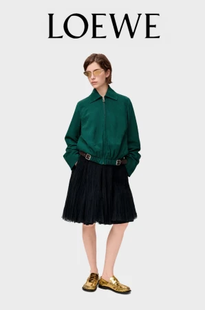 An adult woman poses in a green zip-up jacket, a black pleated skirt with a brown belt, and gold shoes on a grey backdrop.