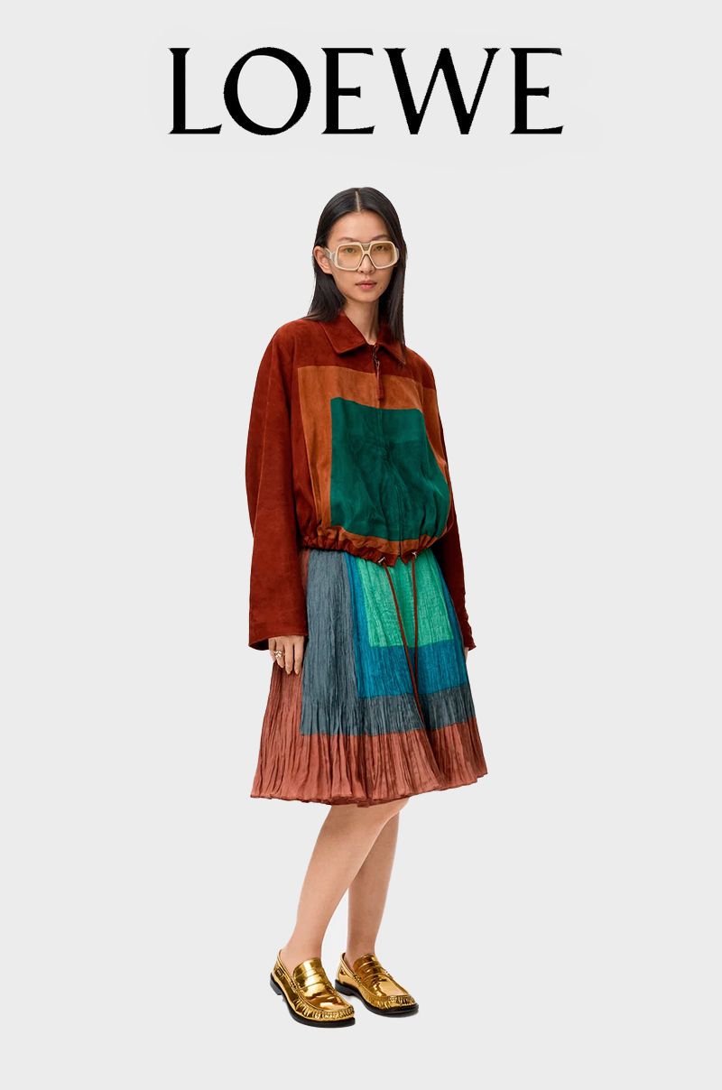 A woman in a patterned dress and gold shoes is posing in a fashion studio, under the Loewe logo. The shot emphasizes her unique style.