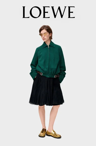 Model wearing a dark green zip jacket, black pleated skirt, and metallic gold shoes against a studio backdrop for Loewe FW25.
