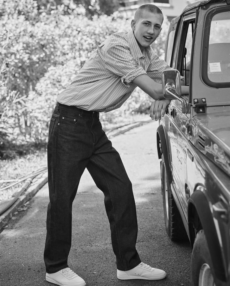 A young adult man leans against a dark SUV, wearing a striped shirt, dark jeans, and white sneakers, in a black and white outdoor setting.
