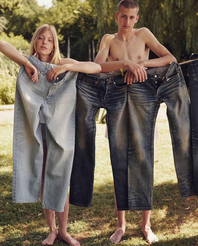 Two young adults pose outdoors with denim jeans hanging from a clothesline, set against a backdrop of trees and natural light.