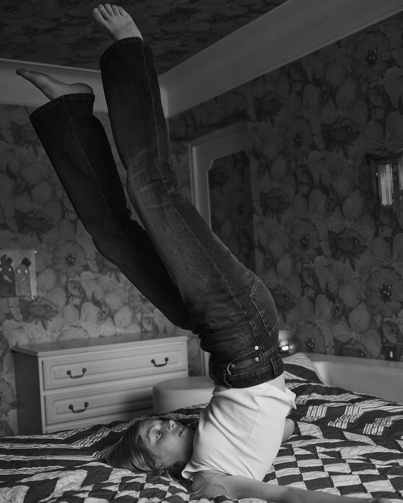 A woman in jeans and a white shirt does a shoulder stand on a bed with a black and white patterned quilt in a bedroom.