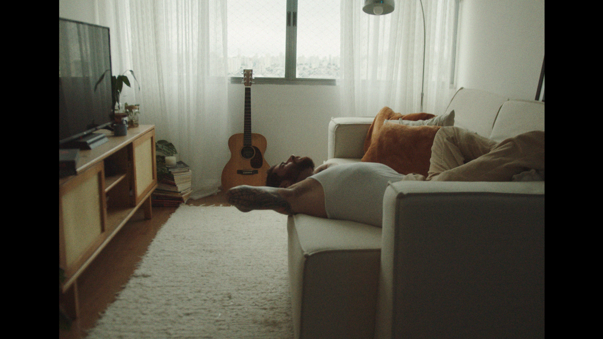 Adult male with beard and tattoos lying on a white couch in a sunlit living room with a guitar in the background.