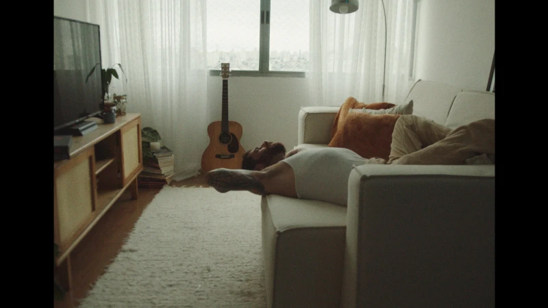 Adult male with beard and tattoos lying on a white couch in a sunlit living room with a guitar in the background.