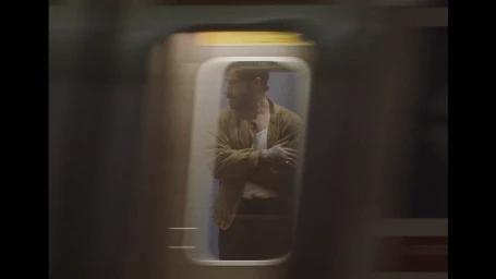 Man in a tan jacket standing inside a subway car seen through a moving train window.