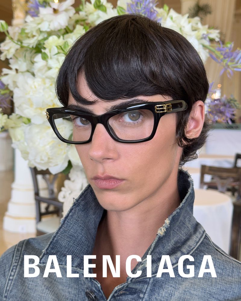Close-up of a person wearing black-framed glasses and a denim jacket, posing in front of flowers. Fashion editorial style.