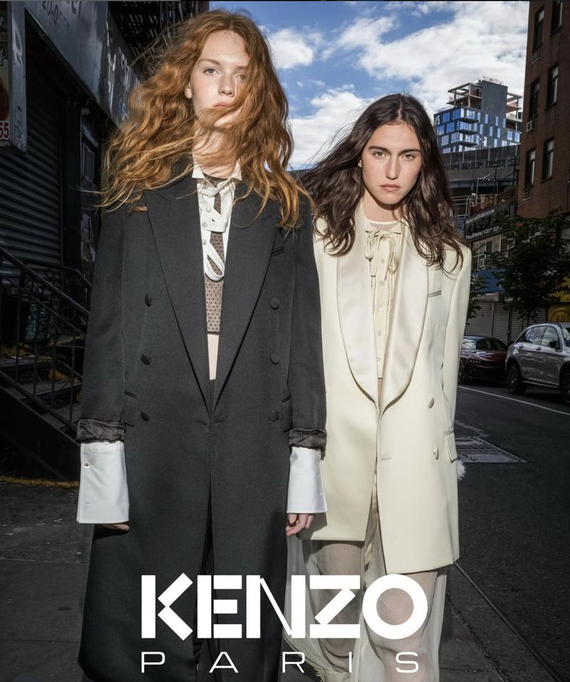 Two young women in stylish blazers stand on a city street for a fashion brand advertisement, featuring an urban backdrop and logo.