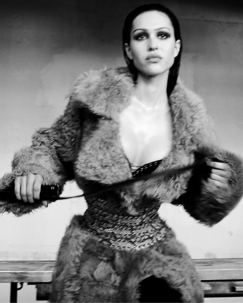 Monochrome portrait of a woman in a fur coat and ornate corset, holding a whip, against a blurred background, in a fashion-forward composition.