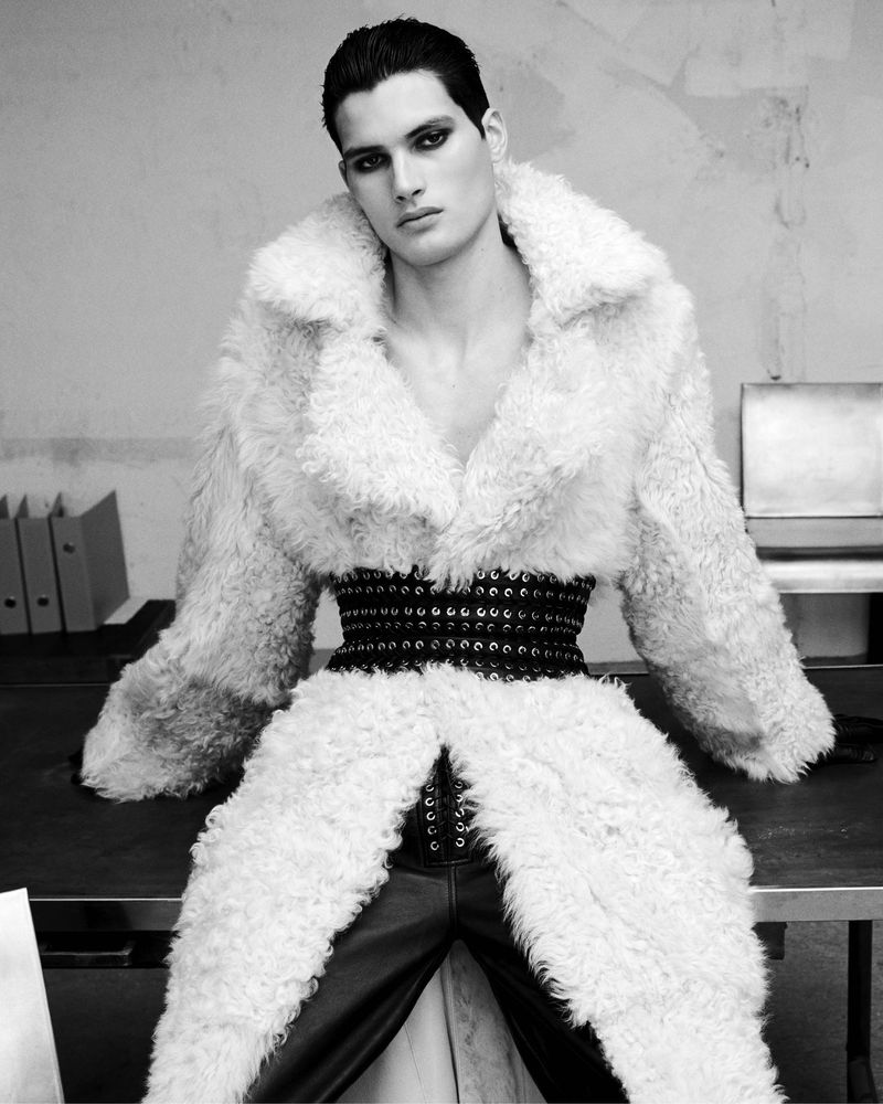 Monochrome fashion shot of a man with dark hair and dramatic eye makeup wearing a white fur coat and leather pants while posing indoors.