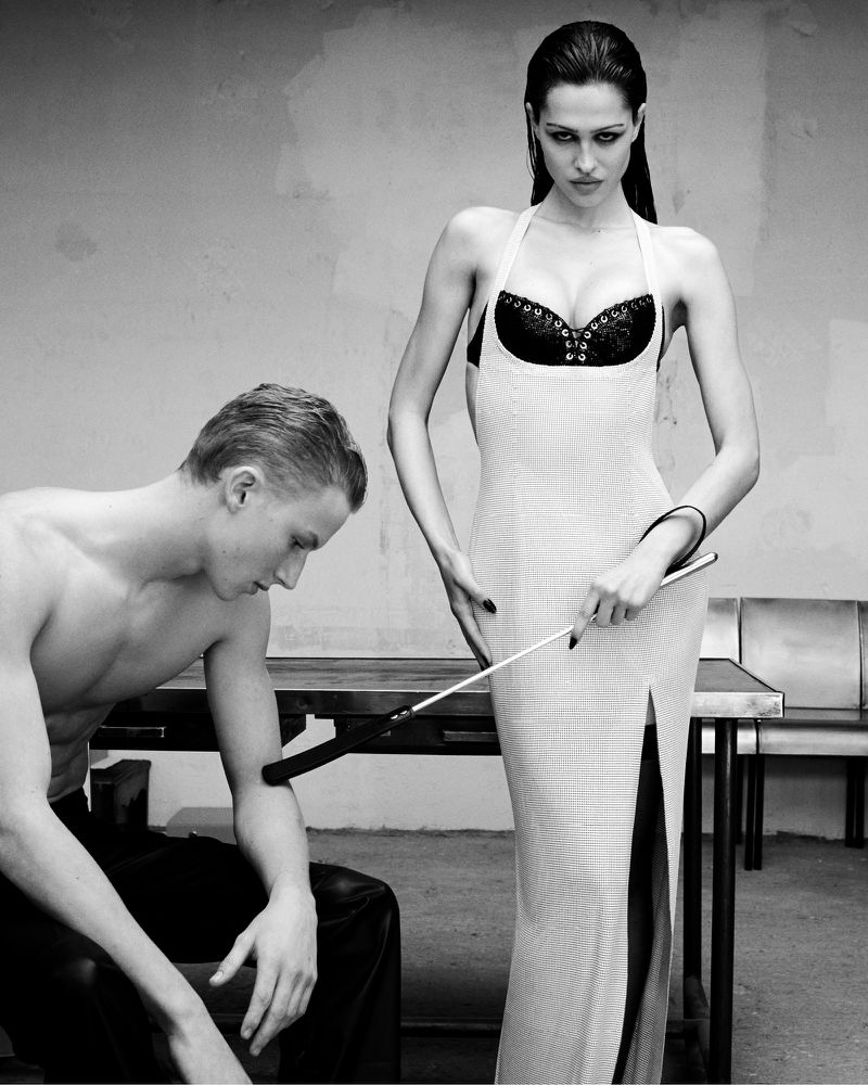 Black and white studio portrait of a woman in a dress holding a riding crop, standing over a shirtless man sitting on a bench.
