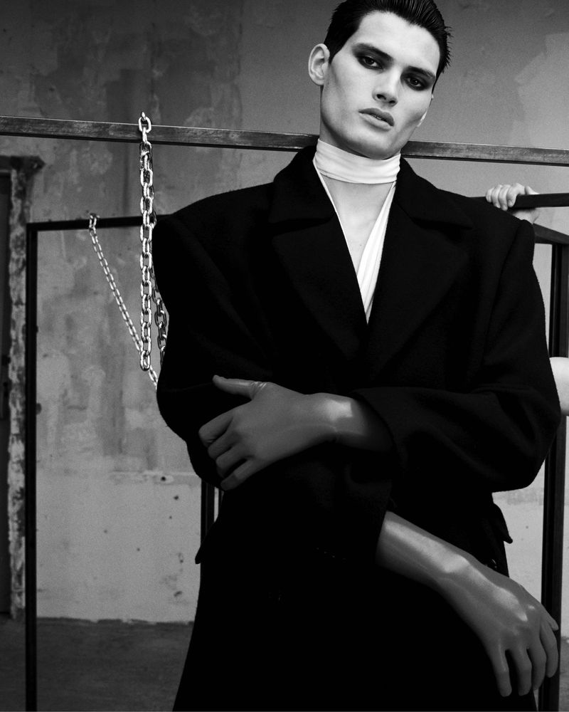 Monochrome portrait of a man in a black coat and white scarf, posing indoors with crossed arms and a neutral expression.