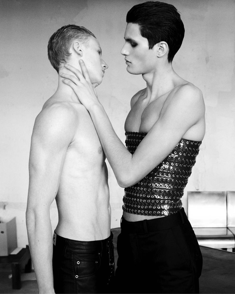 Black and white studio shot of two men, one topless in leather pants with a hand on the neck of the other, who wears a studded top.