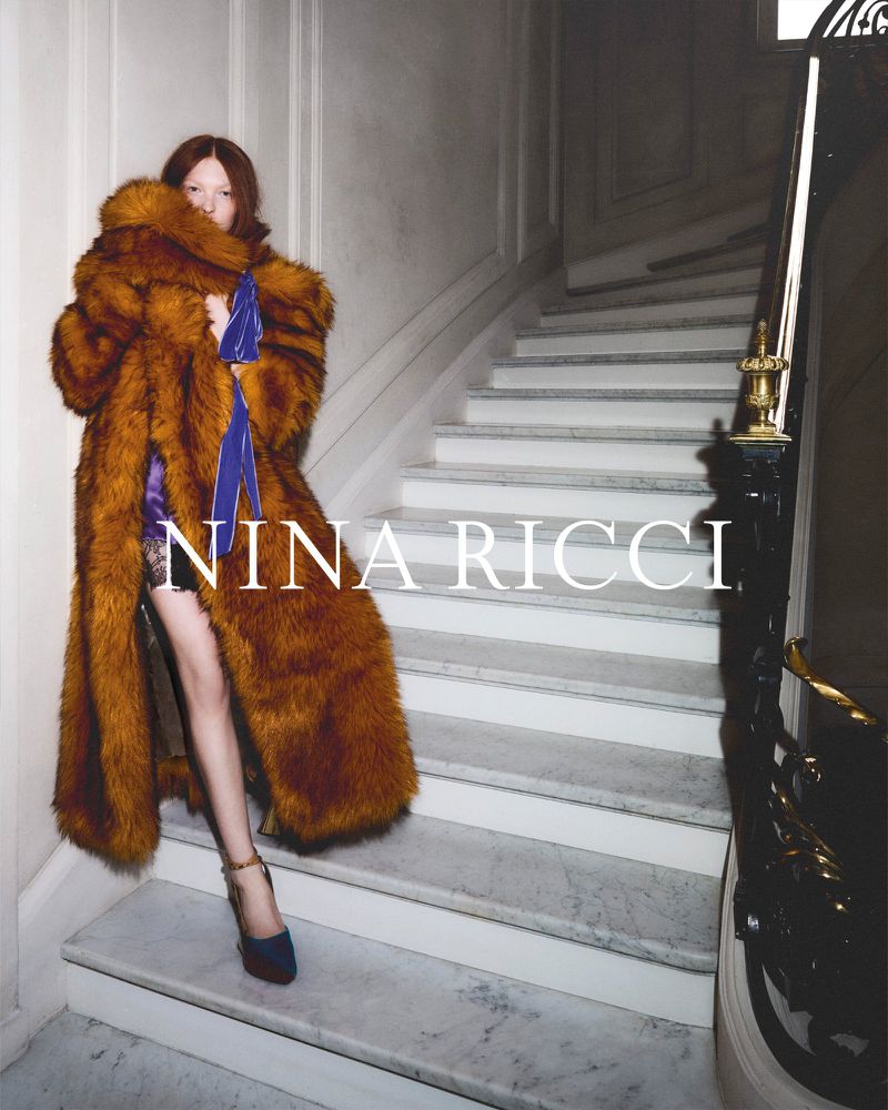 Fashion shot of a model wearing a long brown fur coat and navy high heels, posing on a staircase with a white wall backdrop.