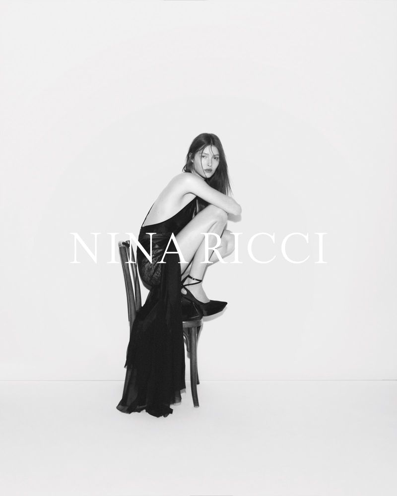 A woman in a black dress sits on a chair in a studio, with the fashion brand Nina Ricci text overlayed on the bright white background.