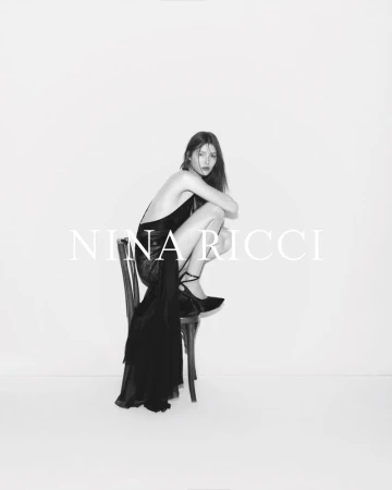 A woman in a black dress sits on a chair in a studio, with the fashion brand Nina Ricci text overlayed on the bright white background.