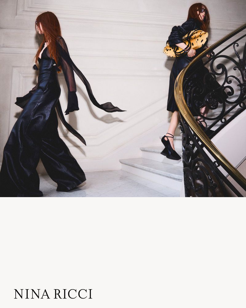 Two fashion models in elegant attire ascend a marble staircase. One carries a textured bag. A Nina Ricci advertisement.