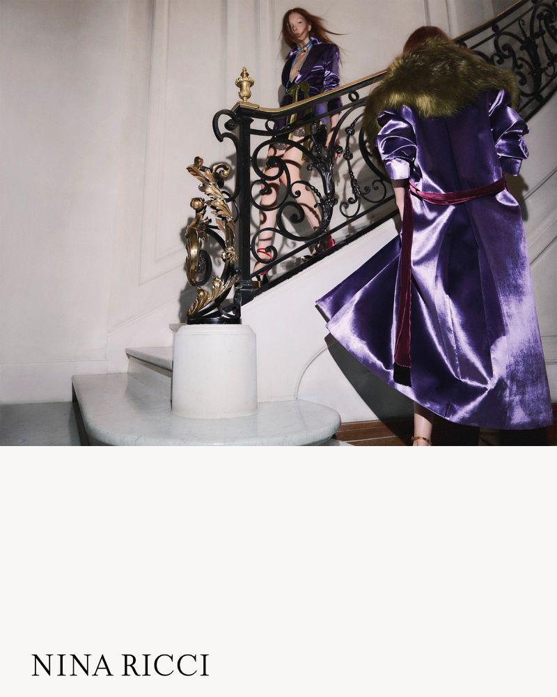 Two fashion models in purple outfits are captured on a white staircase with a decorative black and gold railing in an indoor setting.