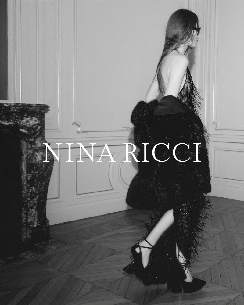 Black and white fashion shot of a woman in a fringed dress and jacket, walking past a marble fireplace, promoting Nina Ricci.