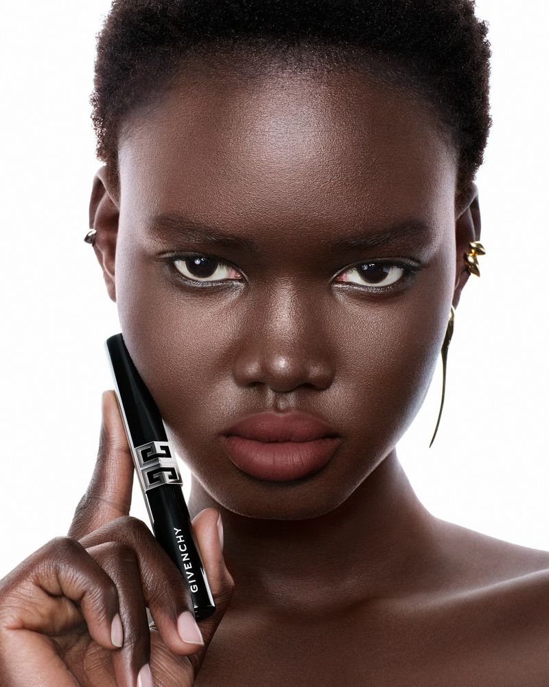 Beauty shot of a young African American woman with dark skin holding Givenchy mascara, wearing gold earrings, against a white background.