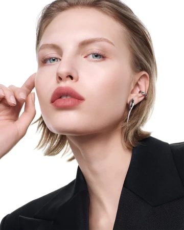 Close-up portrait of a young woman with blond hair, wearing a black blazer and silver earrings against a white background.