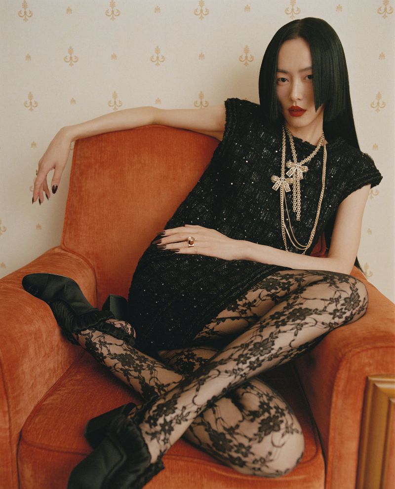A glamorous woman with a blunt bob poses in a sparkling black dress, sheer floral lace stockings, and black shoes while sitting in an orange armchair.