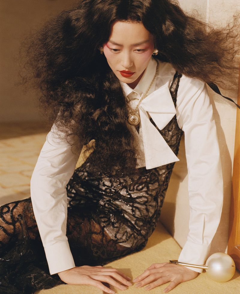 Elegant Asian woman in a lace dress and white shirt poses indoors with dramatic eye makeup and voluminous dark hair, creating a sophisticated fashion image.