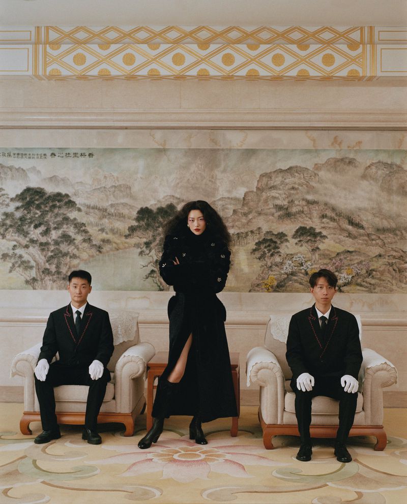 Fashion photo features a confident woman in a black coat flanked by two seated men in formal attire, set against an ornate interior backdrop.