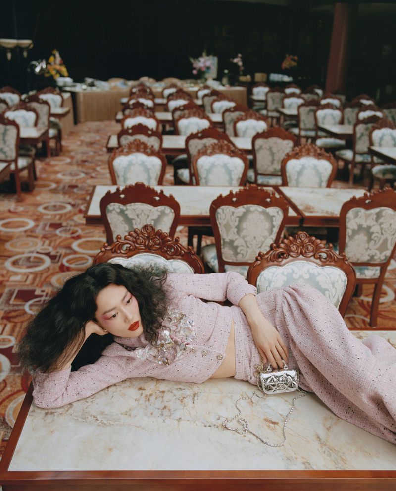 Elegant woman in a pink pantsuit and handbag rests on a marble table in a room with rows of empty chairs and tables.