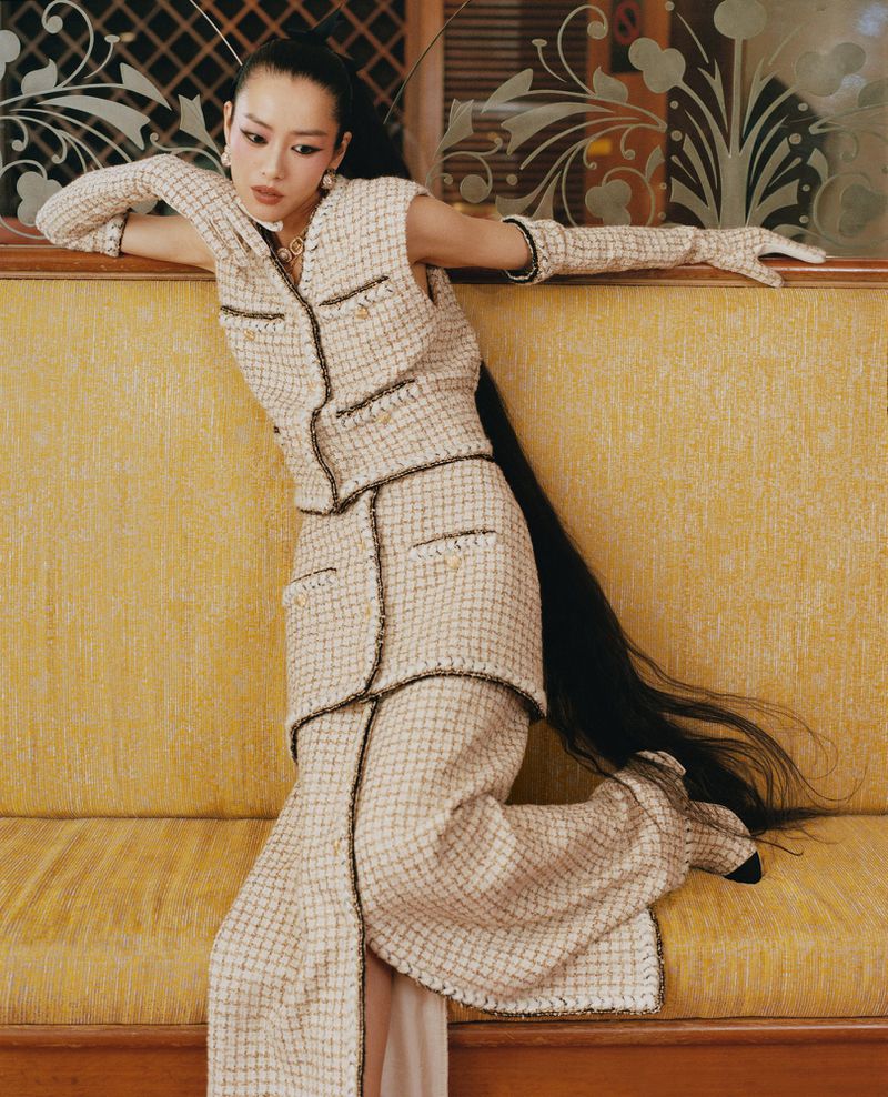 Elegant Asian woman in beige tweed suit with long hair poses on yellow couch in front of floral glass panel, embodying fashion and style.