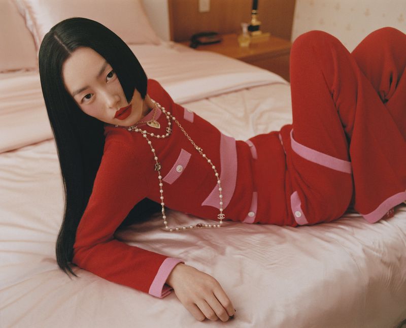 Attractive Asian woman in red outfit lying on a bed in a close-up shot. She has long black hair, red lipstick, and pearl necklace.
