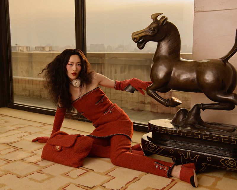 Fashion shot of Asian woman in a red tweed suit sitting near a bronze horse statue, bag at her side, indoor setting with city view.