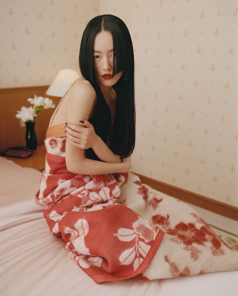 Portrait of a young Asian woman with dark hair wrapped in a floral blanket sitting on a bed in a bedroom with wallpaper.