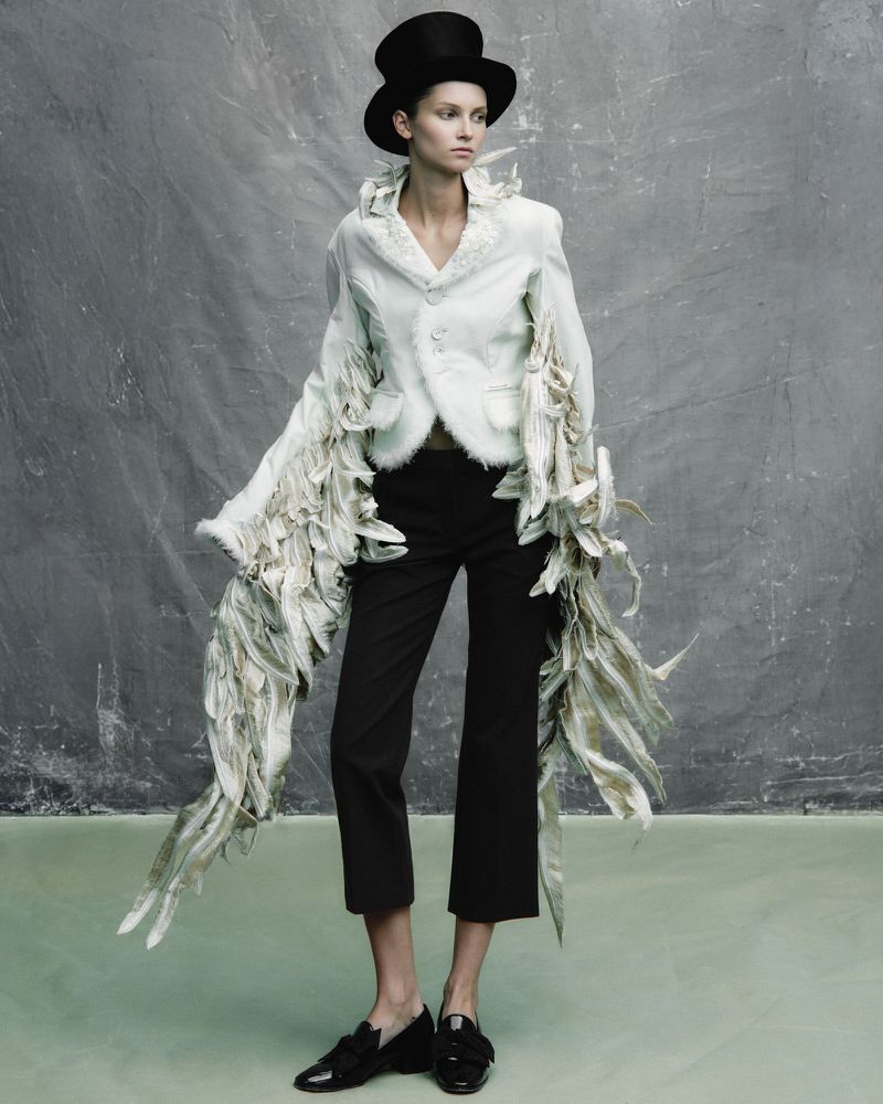 A woman in a feather-trimmed jacket and top hat poses against a mottled gray backdrop. She is wearing black cropped pants and loafers.
